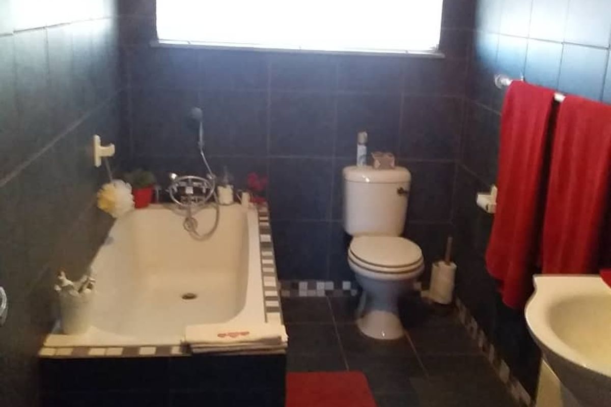 A functional bathroom featuring dark wall and floor tiling, a built-in bathtub with shower mixer, a standard toilet, and a vanity basin.