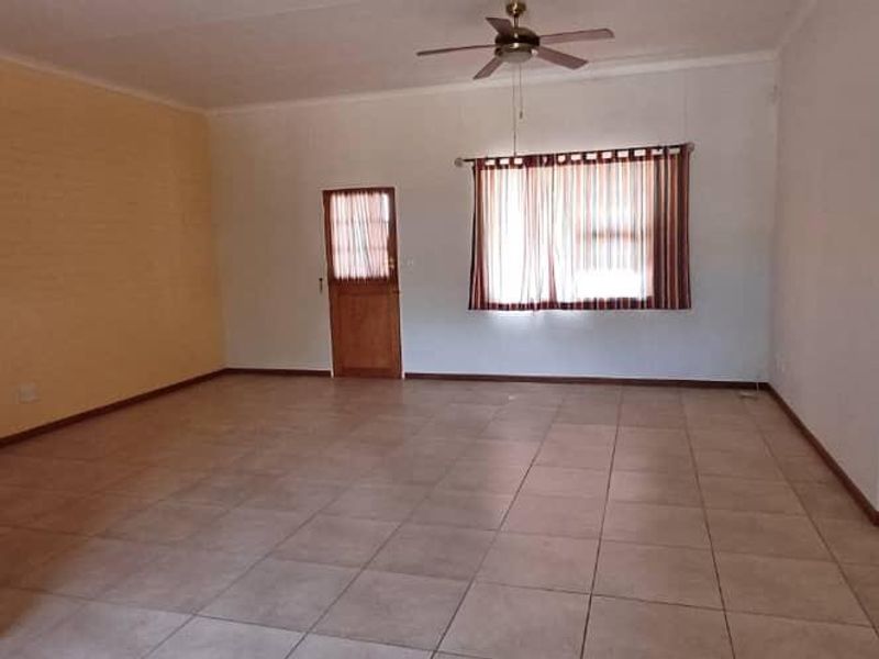A spacious, empty living room featuring neutral tiled flooring, a ceiling fan, and natural light through a window with fitted blinds.