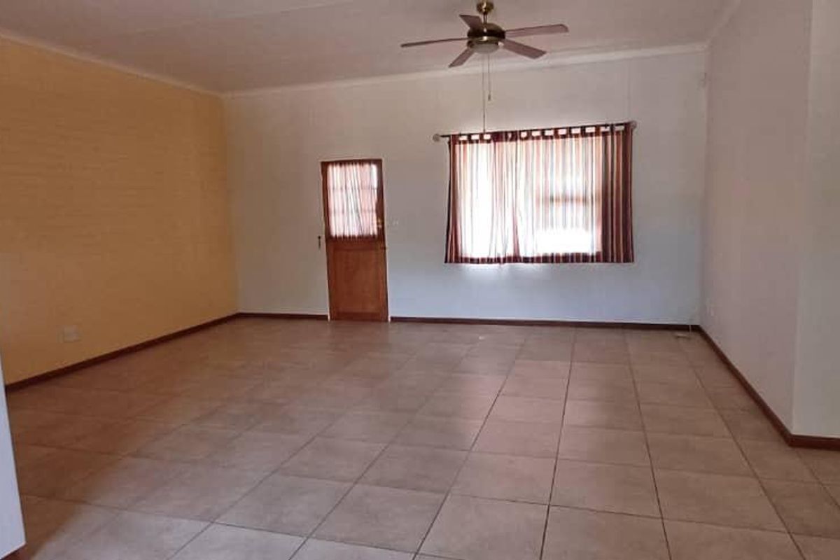 A spacious, empty living room featuring neutral tiled flooring, a ceiling fan, and natural light through a window with fitted blinds.