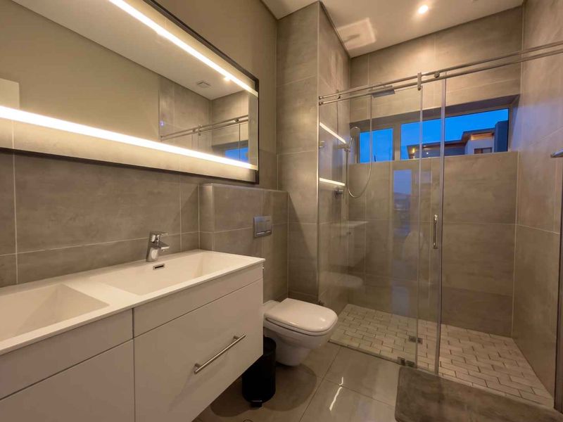 Modern bathroom featuring a double vanity with backlit mirror, wall-mounted toilet, and a spacious glass-enclosed shower with contemporary tiling.