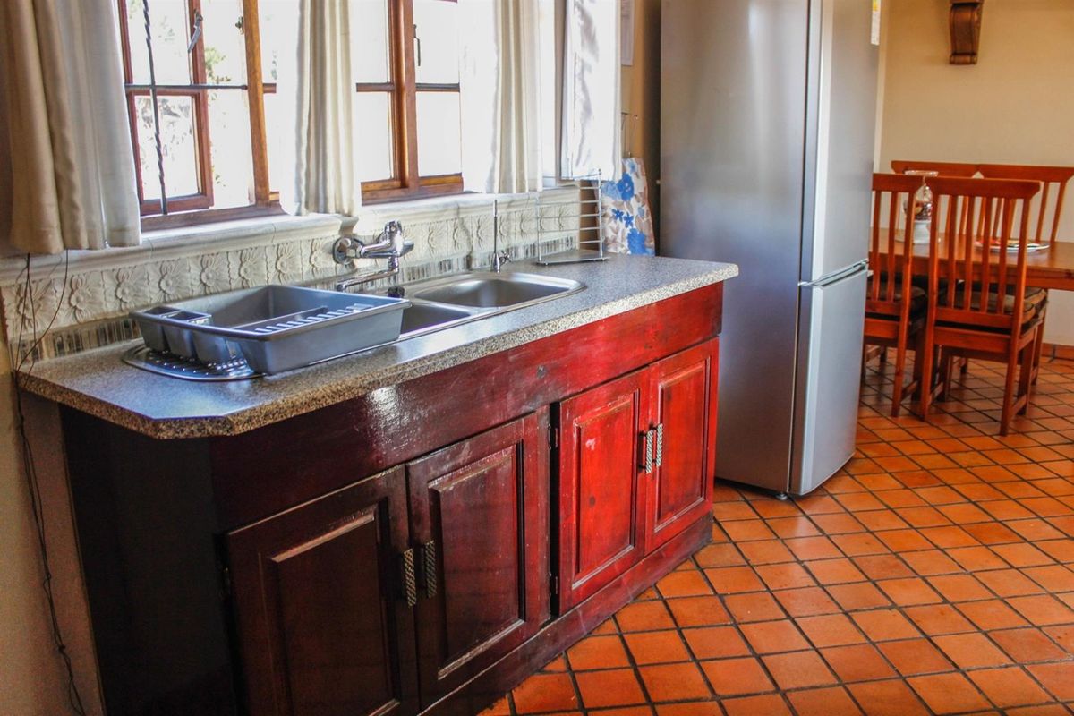 A functional kitchen area featuring wooden cabinetry, granite countertops, a double basin sink, and traditional terracotta floor tiles.