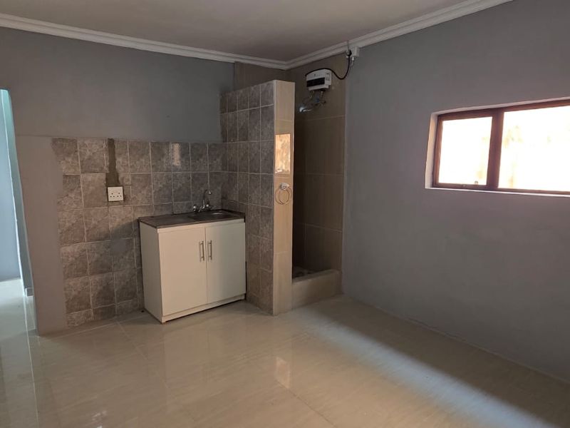 A compact, empty kitchen area featuring tiled flooring, a small sink unit with tiled splashback, and an exposed electric water heater.