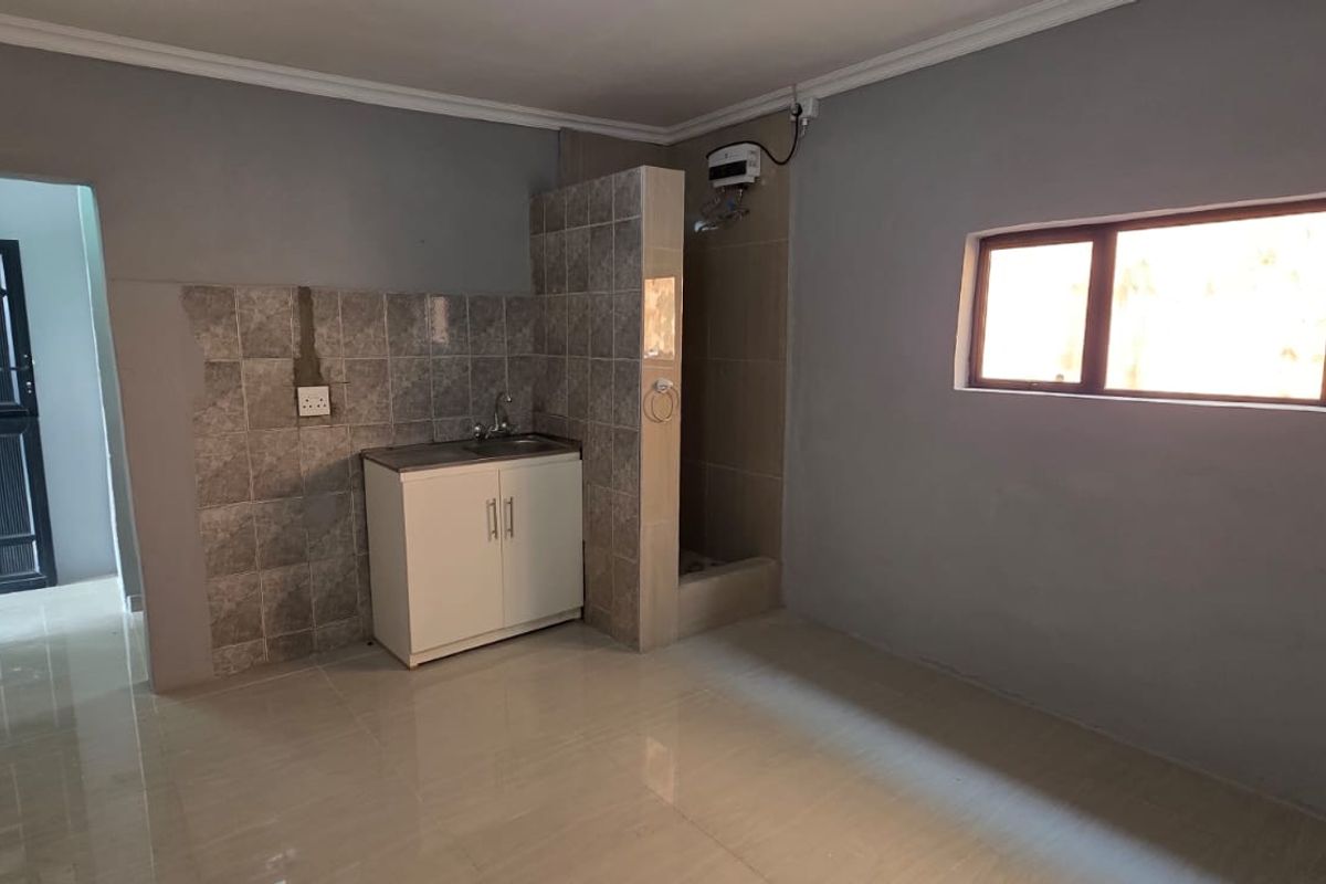 A compact, empty kitchen area featuring tiled flooring, a small sink unit with tiled splashback, and an exposed electric water heater.
