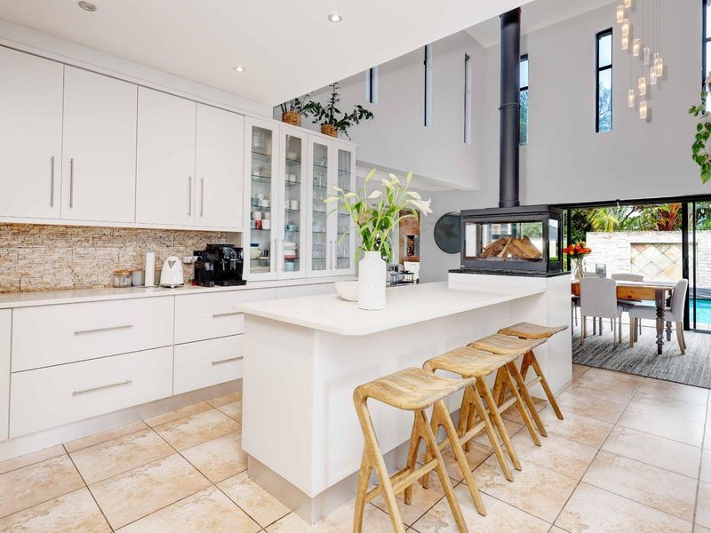 Modern open-plan kitchen featuring sleek white cabinetry, stone countertops, a central island, and a striking double-sided fireplace, seamlessly connecting to the outdoor patio area.