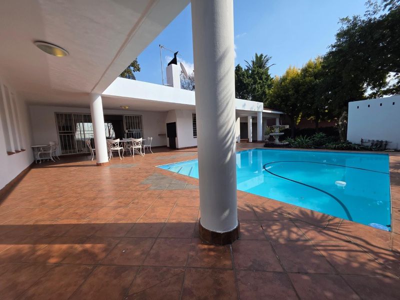 Spacious covered patio area overlooking a swimming pool, featuring tiled flooring, structural columns, and a built-in outdoor fireplace.