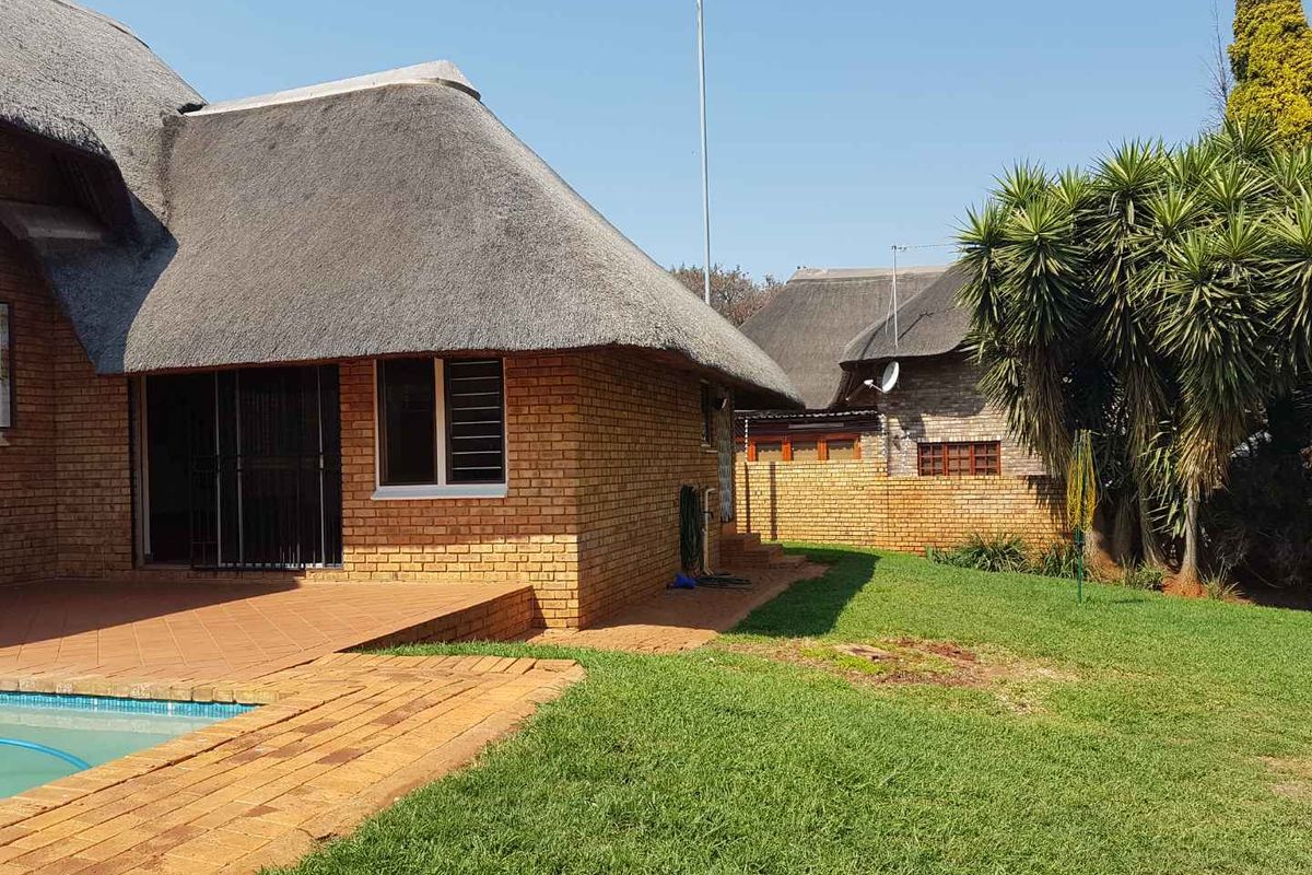 Rear view of a face-brick house featuring a traditional thatched roof, a paved patio area, and a private swimming pool set in a garden.