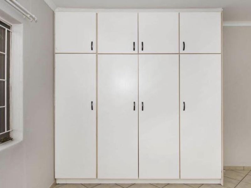 A clean, empty bedroom featuring white built-in cupboards, tiled flooring, and a window fitted with security bars.