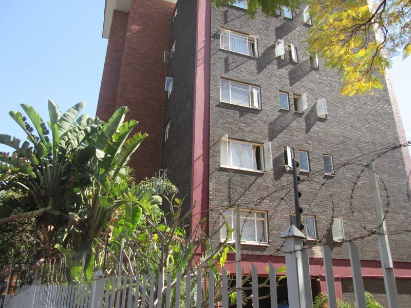 Exterior view of a multi-story apartment building featuring a brick facade, casement windows, and secure perimeter fencing.