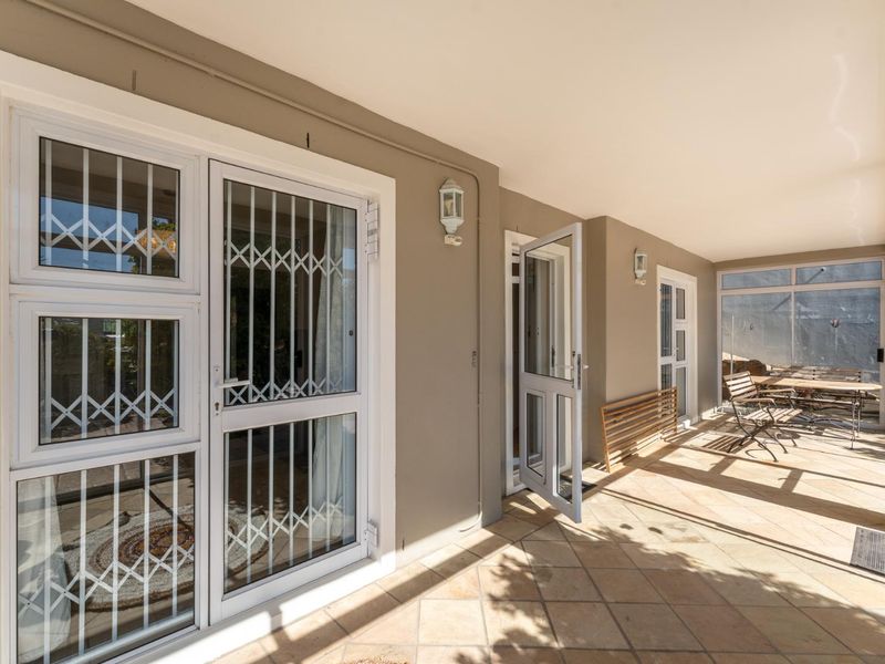A spacious covered patio featuring tiled flooring, secure white-framed doors with security gates, and wall-mounted lighting, providing a functional outdoor living area.