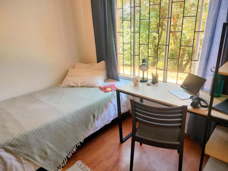 A compact bedroom featuring laminate flooring and a large window fitted with security bars, providing natural light and a view of the outdoors.