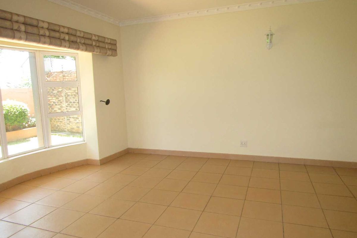 A spacious, empty living room featuring neutral ceramic floor tiles, crown molding, and a large window with a fitted roman blind.