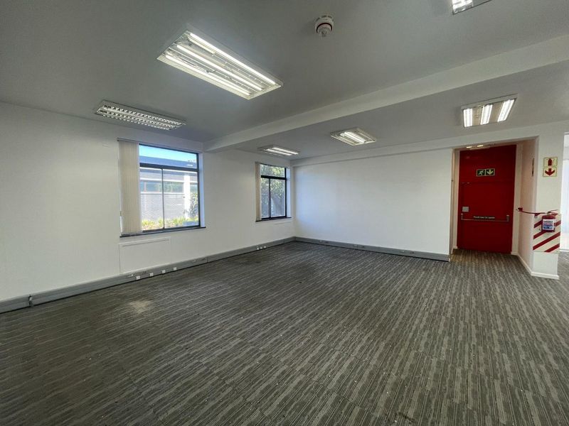 A vacant, open-plan office space featuring neutral walls, carpeted flooring, recessed fluorescent lighting, and a prominent red fire exit door.