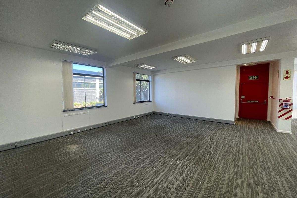 A vacant, open-plan office space featuring neutral walls, carpeted flooring, recessed fluorescent lighting, and a prominent red fire exit door.