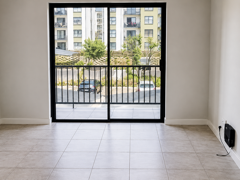 A bright, empty living area featuring neutral tiled flooring and a large sliding glass door leading to a balcony with an urban view.
