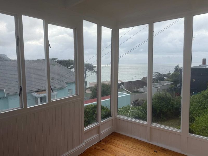 A bright, enclosed sunroom or balcony featuring timber flooring, white paneling, and expansive windows offering scenic coastal views.