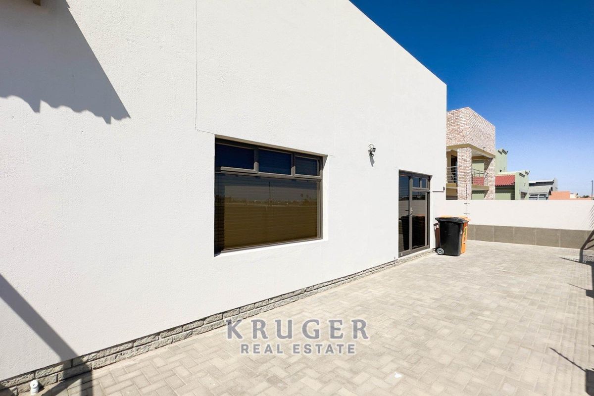 A clean rear exterior view featuring paved patio area, modern white facade, and dark-framed windows and doors.