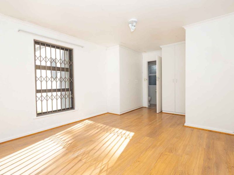 A bright, empty bedroom featuring laminate flooring, built-in cupboards, and security bars on the window, with an adjacent en-suite bathroom.