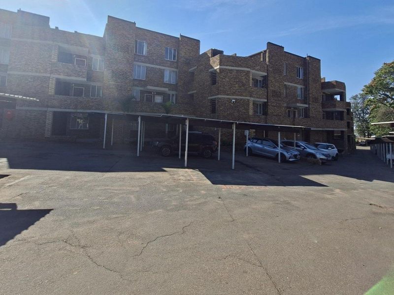 Exterior view of a multi-story brick apartment building featuring covered parking bays and an asphalt driveway.