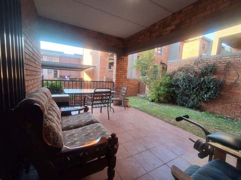 A covered outdoor patio featuring exposed brick walls and tiled flooring, overlooking a small private garden area.