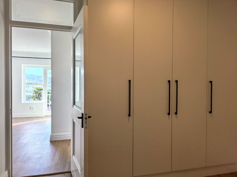 A bright bedroom featuring modern built-in cupboards with sleek black handles and warm timber flooring, leading into an adjacent living space.
