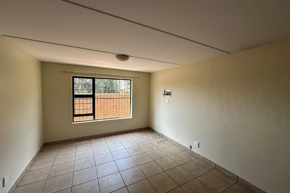 A vacant living room featuring tiled flooring, a large window with security bars, and neutral wall finishes.