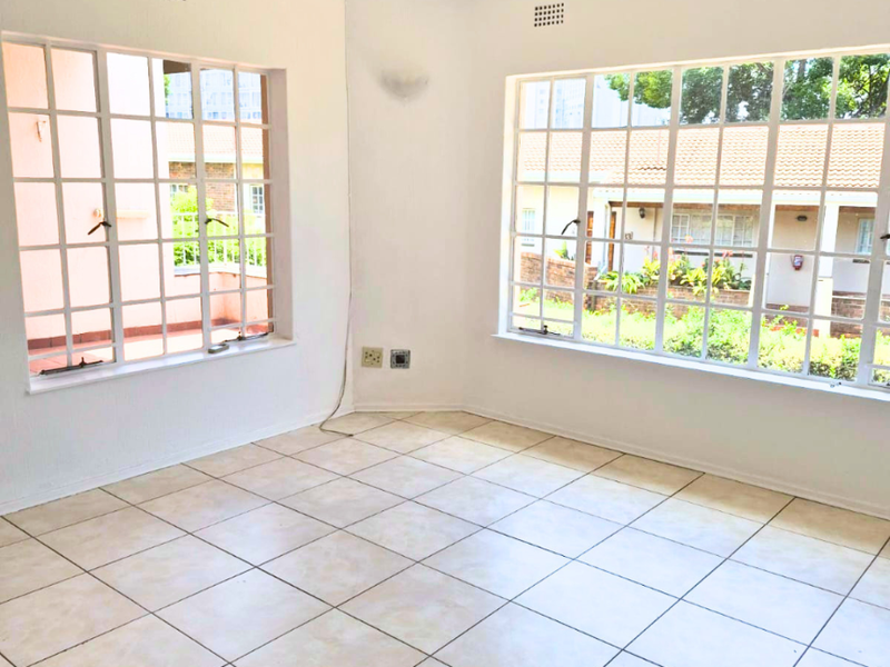 A bright, empty living area featuring ceramic floor tiles, a ceiling fan, and large grid-style windows providing natural light and garden views.