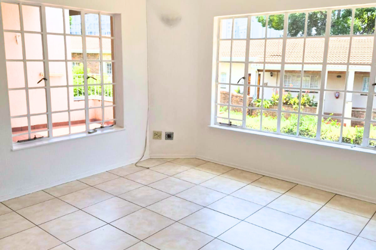 A bright, empty living area featuring ceramic floor tiles, a ceiling fan, and large grid-style windows providing natural light and garden views.