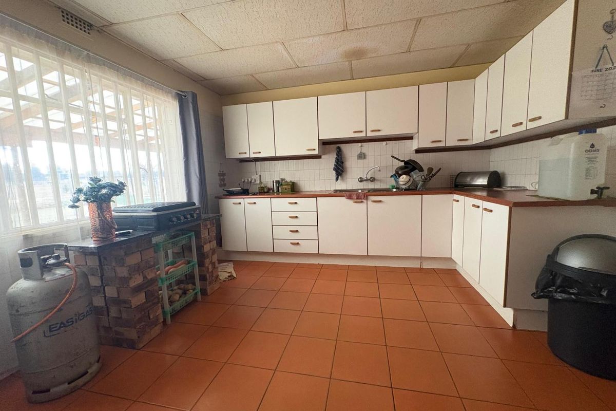 A dated kitchen featuring white cabinetry, tiled countertops, and terracotta floor tiles, with a brick-based prep area and large window.