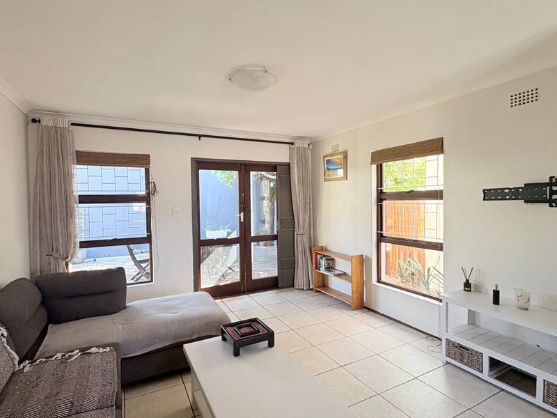 A bright living room featuring tiled flooring, large windows with security bars, and double doors providing direct access to the outdoor area.
