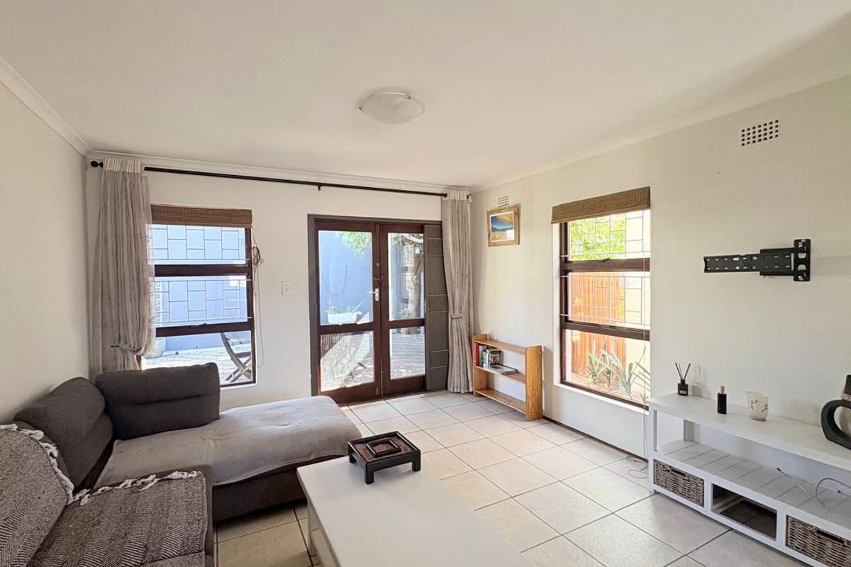 A bright living room featuring tiled flooring, large windows with security bars, and double doors providing direct access to the outdoor area.