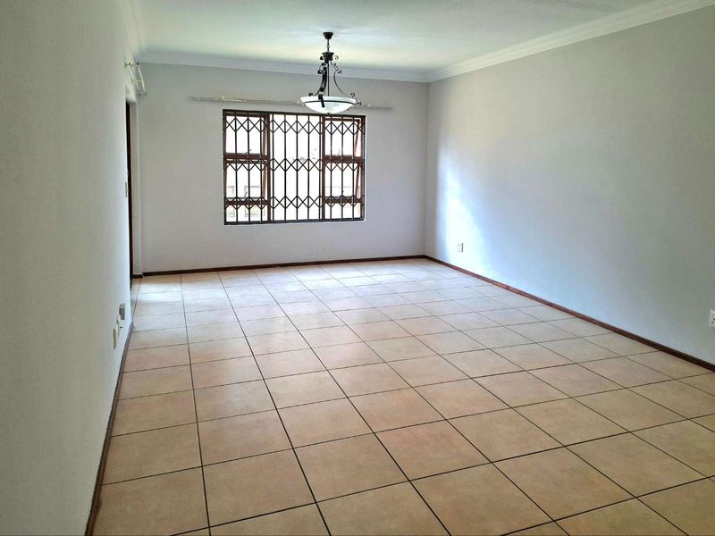 A spacious, empty living room featuring neutral tiled flooring, coved ceilings, and a window fitted with security bars.