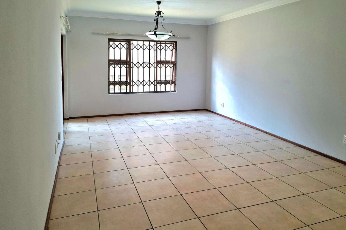 A spacious, empty living room featuring neutral tiled flooring, coved ceilings, and a window fitted with security bars.