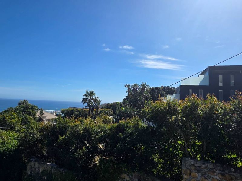 A scenic coastal view featuring lush greenery, a stone retaining wall, and a glimpse of the ocean under a clear blue sky.