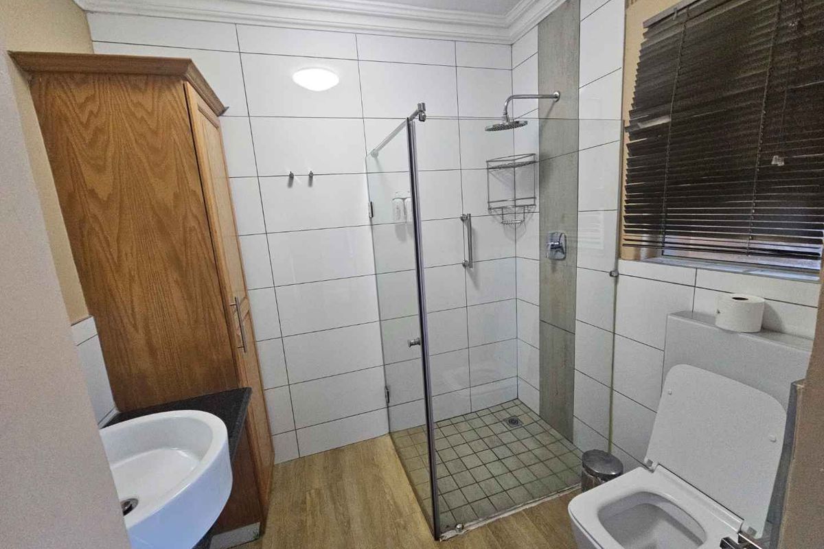 A functional bathroom featuring a glass-enclosed shower, wall-mounted basin, built-in wooden cabinetry, and tiled flooring.