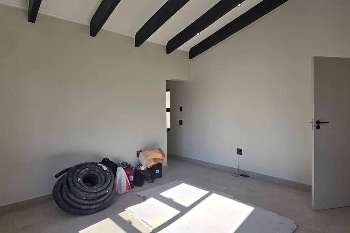A bright, empty bedroom featuring high ceilings with exposed dark timber beams, tiled flooring, and recessed lighting.