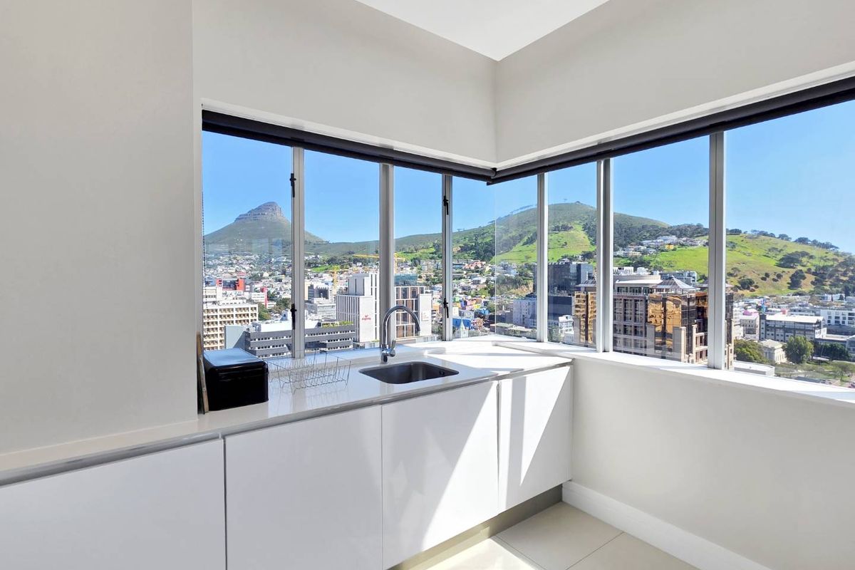 A modern, minimalist kitchen area featuring white gloss cabinetry, stone countertops, and expansive corner windows offering panoramic city and mountain views.