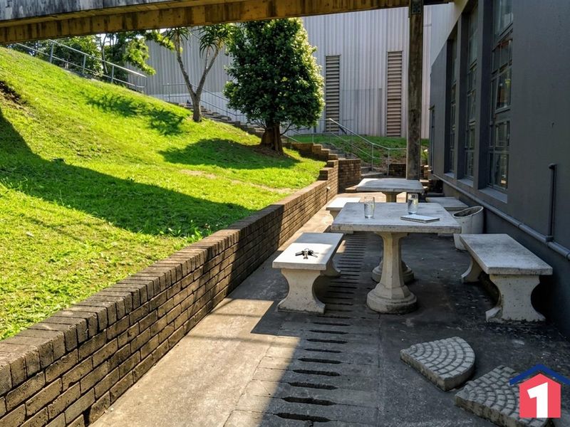 Outdoor patio area featuring fixed concrete tables and benches, set against a brick retaining wall and landscaped embankment within an industrial property setting.