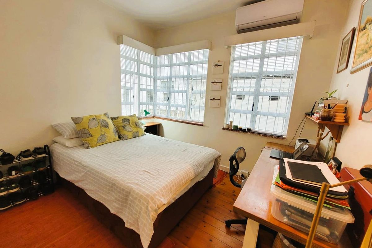 A cozy bedroom featuring polished timber flooring, large corner windows with fitted blinds, and a wall-mounted air conditioning unit.