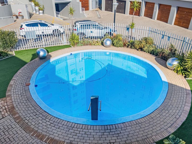 A well-maintained circular swimming pool area featuring paved surrounds, artificial turf, and secure metal perimeter fencing within the complex grounds.