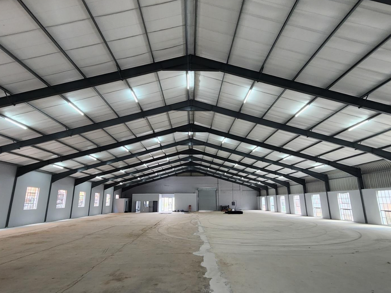 Spacious industrial warehouse featuring a high-clearance steel portal frame, concrete flooring, ample natural light through steel-framed windows, and a large roller shutter door.