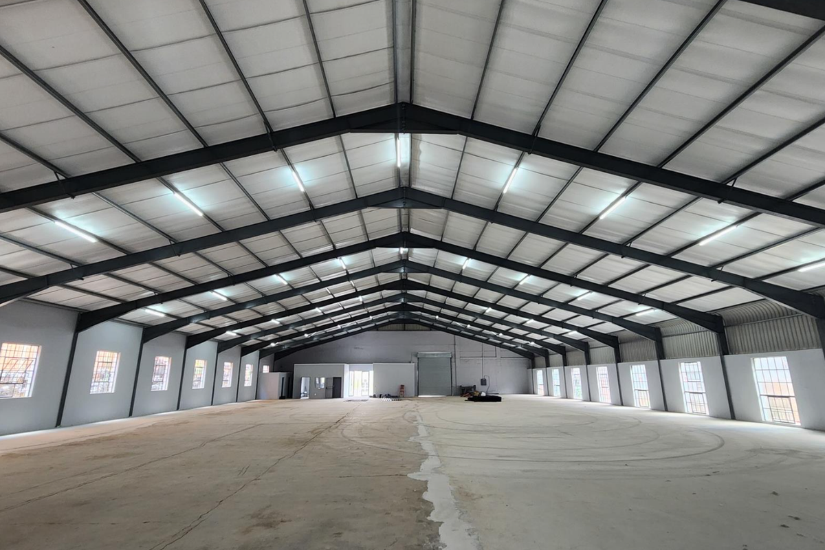 Spacious industrial warehouse featuring a high-clearance steel portal frame, concrete flooring, ample natural light through steel-framed windows, and a large roller shutter door.