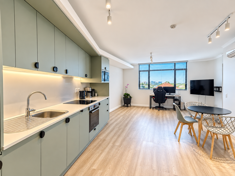 Modern open-plan living area featuring a sleek kitchen with sage green cabinetry, integrated appliances, light wood-look flooring, and large windows with urban views.