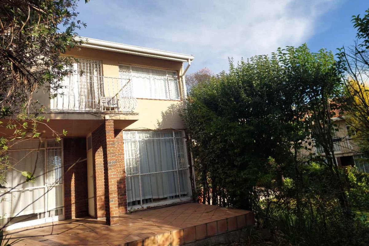 Rear view of the townhouse featuring a tiled patio, upper-level balcony, and established garden surroundings with brick and plaster facade.
