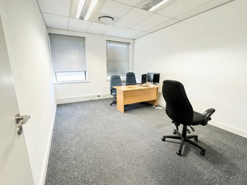 A functional office space featuring carpeted flooring, recessed ceiling lighting, and dual windows with fitted venetian blinds.