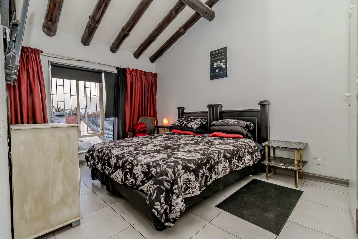 A bedroom featuring exposed timber ceiling beams, tiled flooring, and a window with security bars, offering a rustic architectural aesthetic.