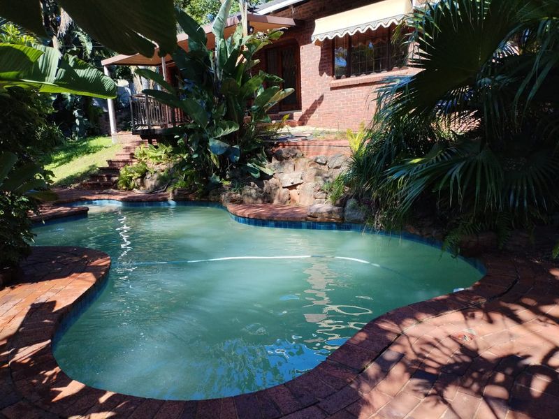 A backyard swimming pool area featuring brick paving, a stone retaining wall, and a brick house facade with an awning.