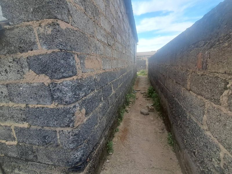 A narrow side passage between a concrete block house wall and a boundary wall, showing signs of wear and unfinished ground surfaces.