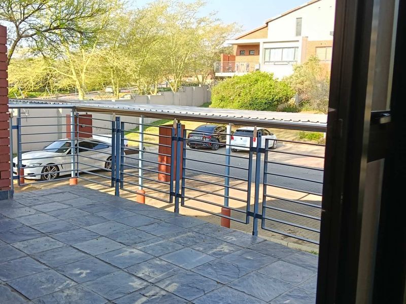 A covered balcony featuring slate tile flooring, a metal balustrade, and a brick feature wall, overlooking the complex parking area.