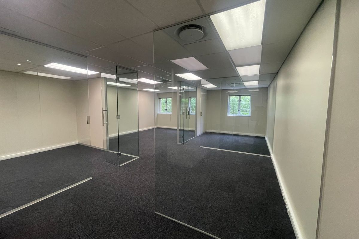 A modern, empty office space featuring glass partitioning, suspended ceilings with recessed lighting, and dark carpeting, offering a clean and professional work environment.
