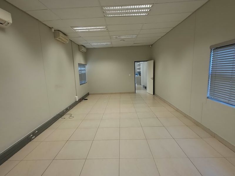 A spacious, empty office area featuring tiled flooring, suspended ceilings with recessed lighting, and multiple wall-mounted air conditioning units.